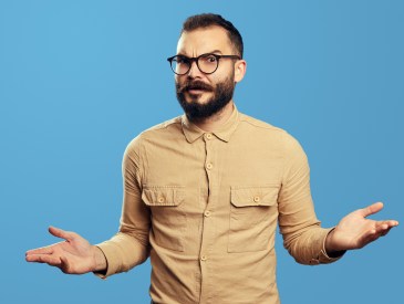 Young bearded man in bewilderment shrugging his shoulders and looking at camera with confusion, isolated over blue background