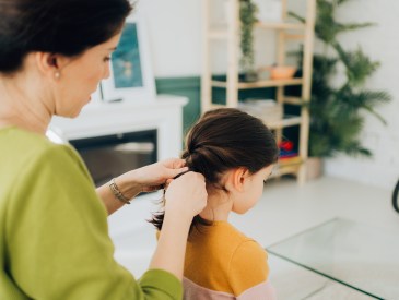 Photo of a mother braiding her daughter's hair in the living room, as they are getting ready for the day.