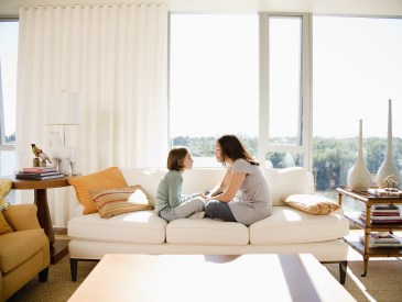 Parent in gray dress sits across from young child in green top and jeans on white sofa in living room.