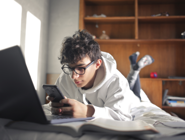 Teen boy looking at his phone while in front of a computer, lying on his bed.